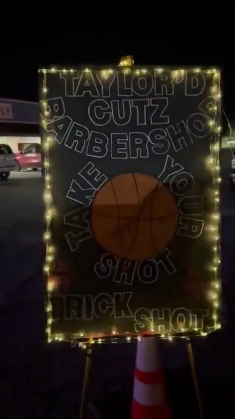 A hand-lettered sign reading Taylor'D Cutz Barbershop, Take Your Shot, Trick Shot, with a basketball in the middle, lit with string lights at night