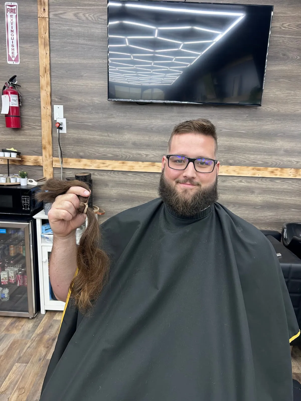 Man with glasses and a full dark beard sitting under a black cape, holding up a length of cut-off hair