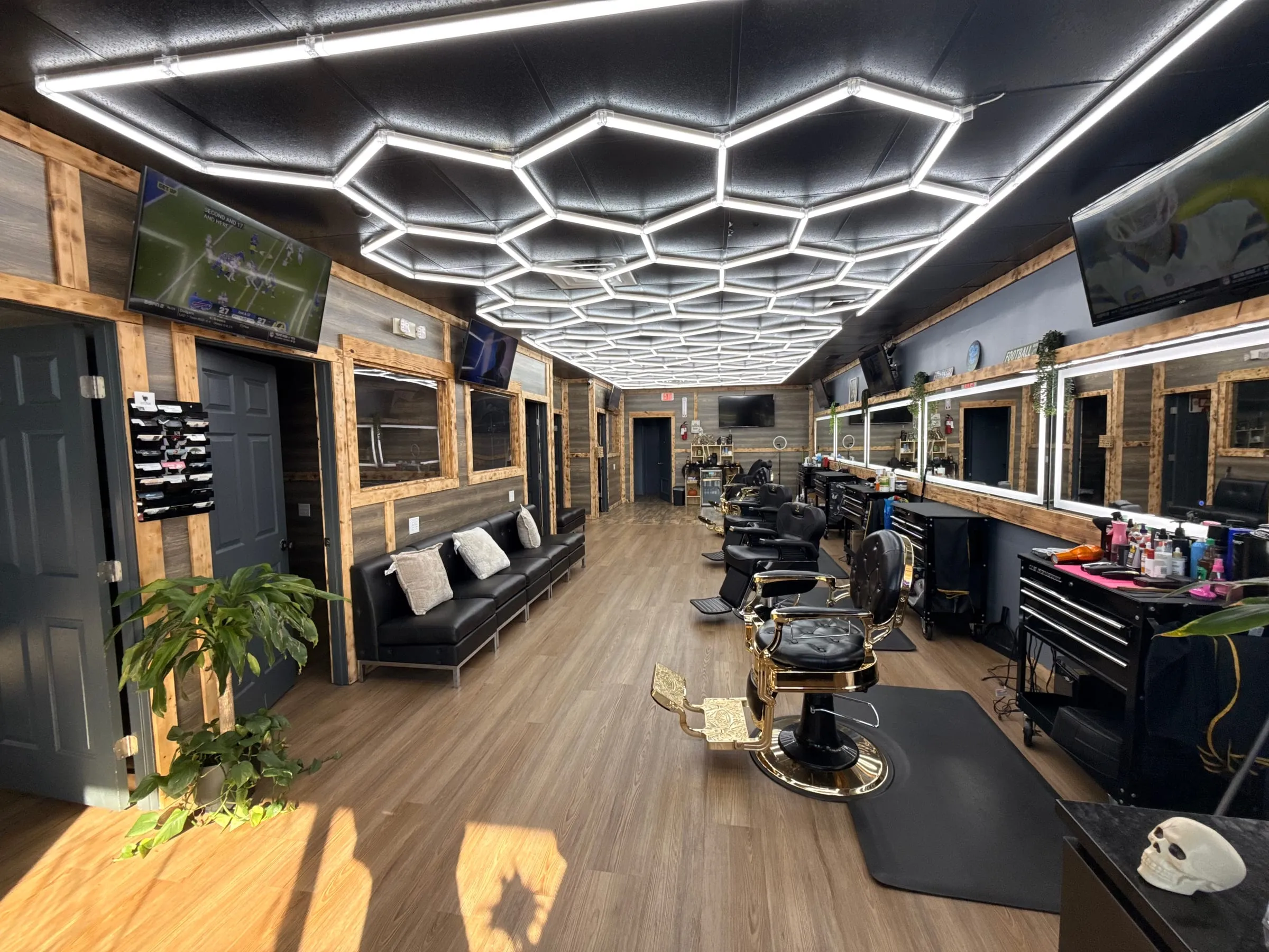 Inside Taylor'D Cutz: a hexagon LED ceiling over a row of black-and-gold barber chairs, lit wood-framed mirrors on the right and a black leather waiting couch on the left.